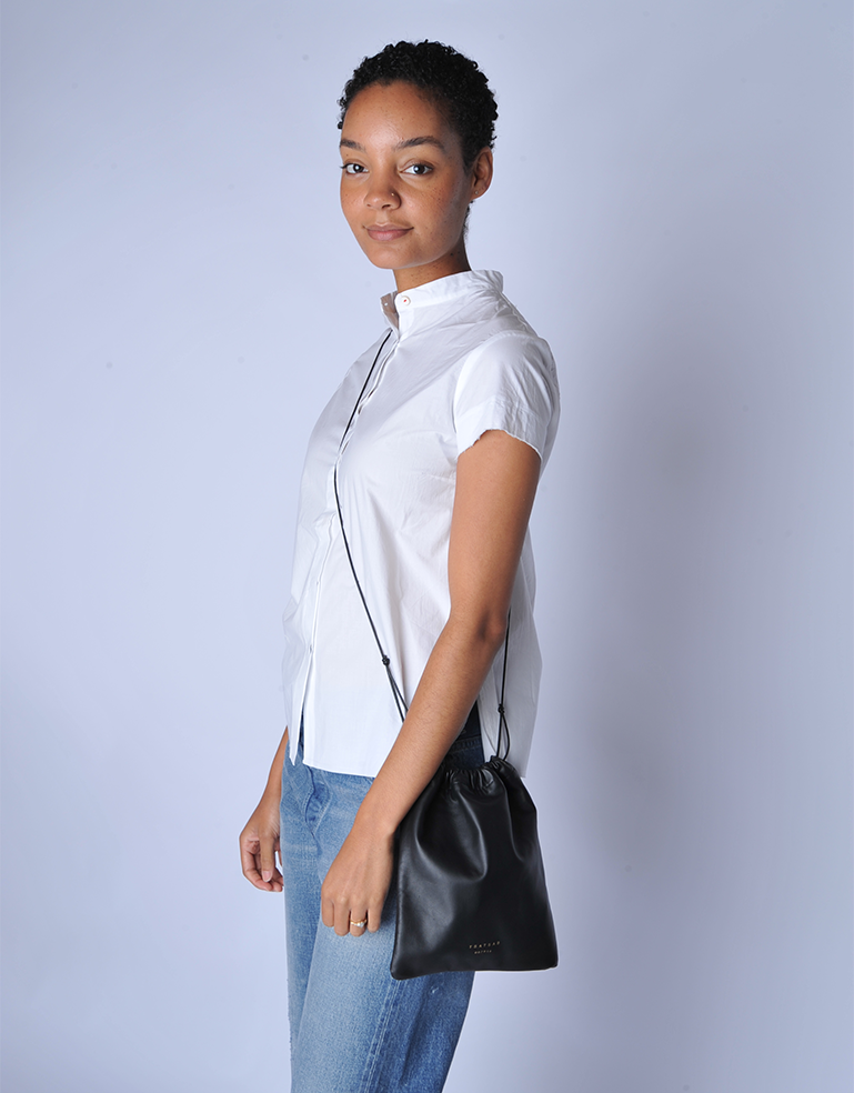 Woman wearing a white shirt and blue jeans, holding a TSATSAS ENA shoulder bag in black lamb nappa leather against a light gray background
