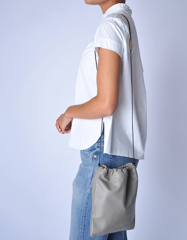 Person wearing a white shirt and blue jeans with TSATSAS ENA shoulder bag in quartz grey lamb nappa leather with drawstring closure detail on a light gray background