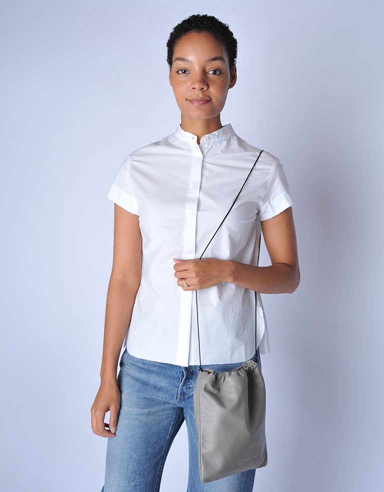 Woman wearing a white shirt and blue jeans, holding a TSATSAS ENA shoulder bag in quartz grey lamb nappa leather with drawstring closure detail against a light background
