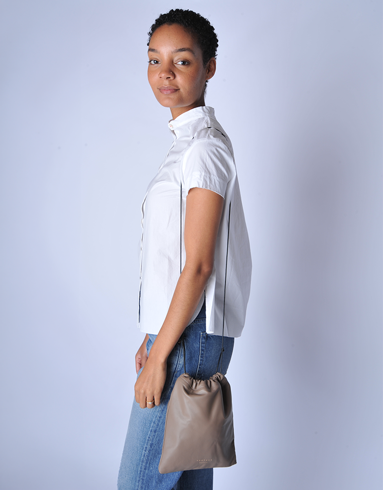 Woman wearing a white blouse and blue jeans, holding a TSATSAS ENA shoulder bag in quartz taupe lamb nappa leather with drawstring closure detail against a plain background
