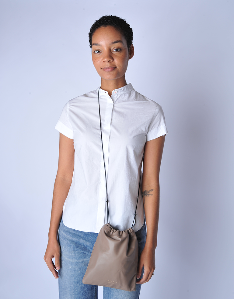 Woman wearing a white short-sleeve shirt holding a TSATSAS ENA bag with solid brass hardware detail on quartz taupe lamb nappa leather against a light gray background