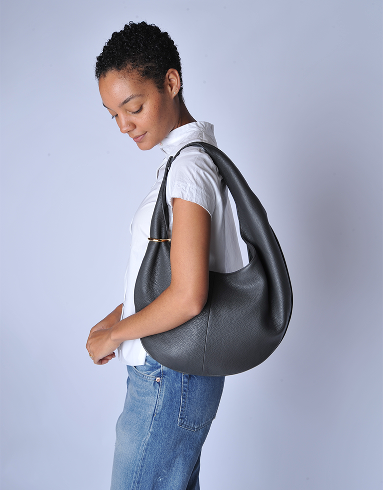 Woman holding a Graphite grey calfskin handbag with an adjustable strap, TSATSAS HALO against a plain background